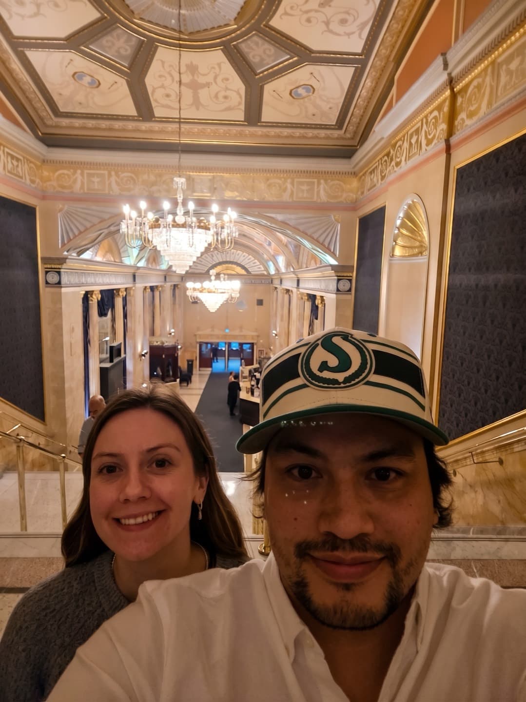 Chelsea and Garnet smiling together beneath the chandeliers of an ornate lobby